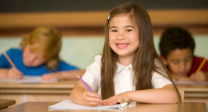 Girl sitting at a desk smiling with a pencil in her hand and two boys writing at desks in the background.